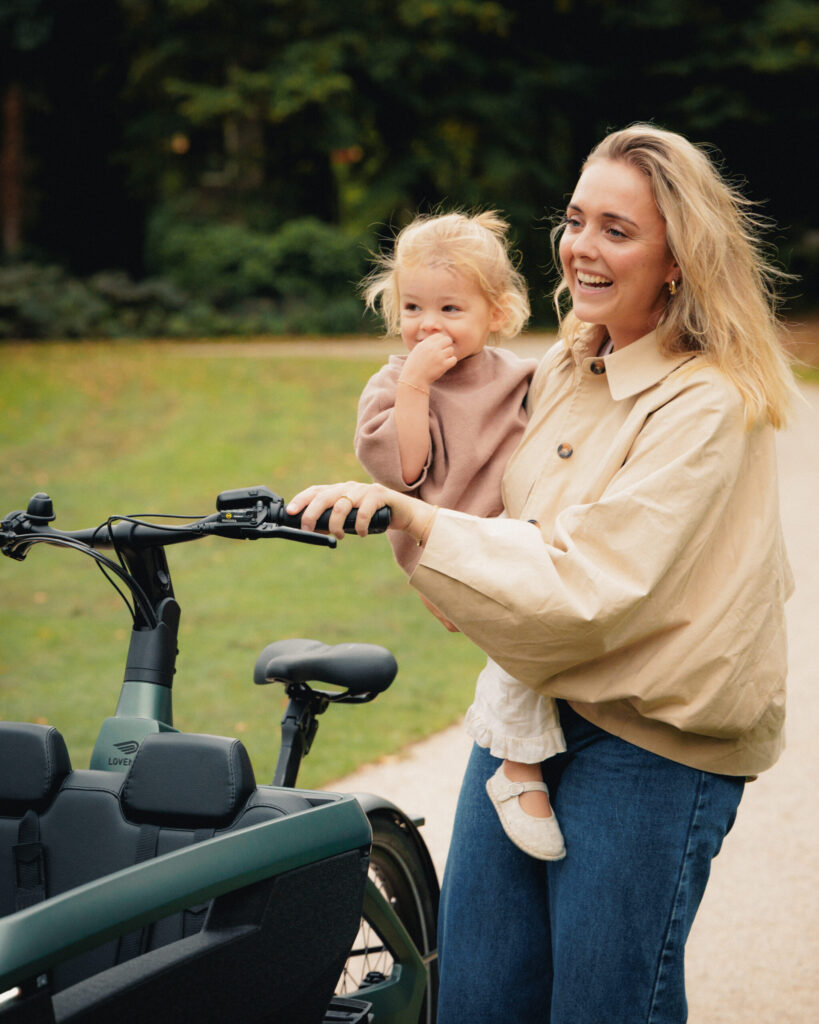 Mother holding a laughing child while standing beside a Avenue green Lovens Explorer 2 cargo bike