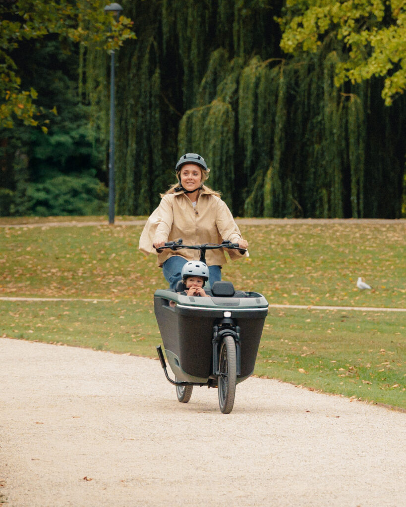 Parent riding a Lovens Explorer 2 cargo bike with a child in the front box through a park